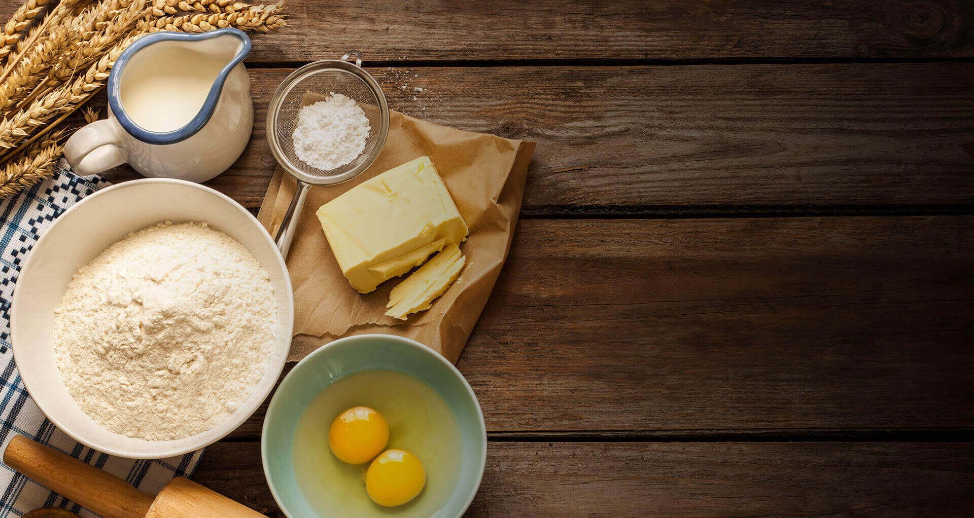 Eine Ansammlung von Backzutaten: Mehl, Butter, Eier, Salz und Milch, fein säuberlich auf einer hölzernen Unterlage angeordnet, mit Weizenhalmen in der Nähe.