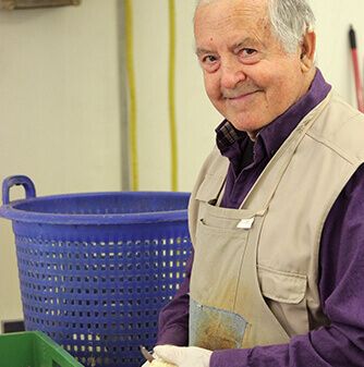 Eine ältere Person in einer beigen Schürze steht neben einem blauen Korb und ist mit der Vorbereitung oder Sortierung von Gegenständen beschäftigt. Im Vordergrund steht eine grüne Tonne.