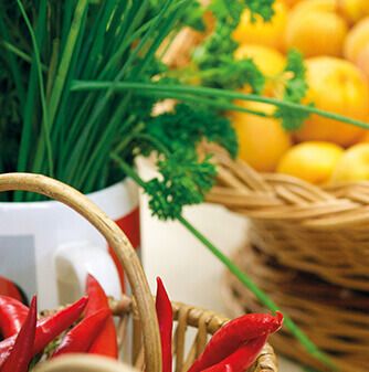 Nahaufnahme eines Weidenkorbs, gefüllt mit roten Chilis, neben einem Korb mit gelben Zitronen und frischen Kräutern wie Schnittlauch und Petersilie.